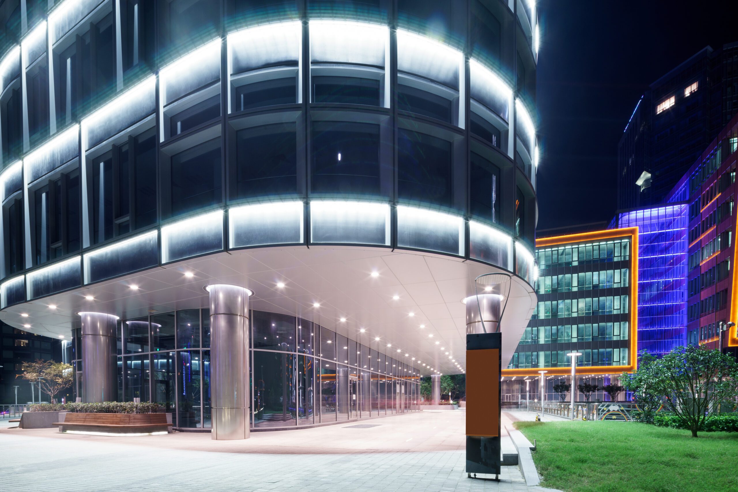 Night view of a modern curved glass building illuminated from within, with a paved walkway, street lamps, and colorful building facades in the background.